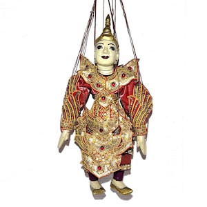 May include: A traditional Burmese marionette puppet. The puppet is dressed in a red and gold costume with intricate detailing, including sequins and jewels. It has a painted face and a golden headdress. The puppet is suspended by strings.