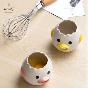 May include: Two white ceramic egg separators shaped like cartoon chicks, with yellow and pink accents. One contains an egg yolk. A stainless steel whisk with a wooden handle is also present. The image is on a light wood surface.