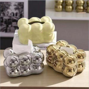 Op de afbeelding: Drie decoratieve tissueboxhouders. Eén is lichtgeel, in de vorm van een wolk, en staat op een boek. Een andere is zilverkleurig en de derde is goudkleurig. Alle drie hebben een bubbelachtig ontwerp en bevatten witte tissues.