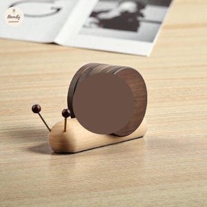 May include: A wooden snail-shaped coaster set. The snail's body is light-coloured wood, with a dark brown shell made of stacked circular coasters. Two small, dark brown spheres form the snail's eyes.