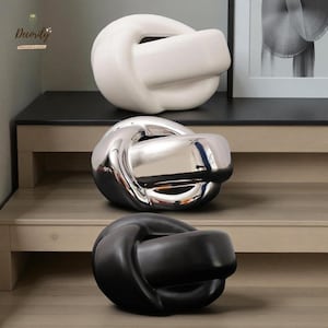 Może przedstawiać: Trzy dekoracyjne rzeźby w kształcie węzłów w kolorze białym, srebrnym i czarnym. Rzeźby są eksponowane na drewnianej powierzchni. Biała rzeźba znajduje się na górze, srebrna pośrodku, a czarna na dole. Rzeźby mają gładkie, błyszczące wykończenie.