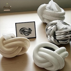 Może przedstawiać: Cztery dekoracyjne ceramiczne pojemniki na chusteczki w kształcie węzła. Uchwyty są w kolorze białym, beżowym i srebrnym. Obecny jest również oprawiony czarno-biały abstrakcyjny nadruk artystyczny.