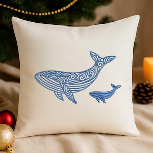 Puede incluir: Cojín cuadrado color crema con un diseño bordado en azul de una ballena grande y otra más pequeña. Las ballenas tienen patrones detallados. El cojín está sobre una superficie neutra con decoraciones navideñas en el fondo.