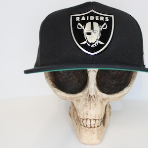 May include: Black baseball cap featuring the Raiders logo in silver. The cap has a flat peak with a green underside. The hat is displayed on a skull.