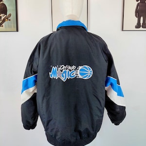 Puede incluir: Chaqueta de baloncesto Orlando Magic negra vintage. La chaqueta presenta un cuello azul, rayas azules y blancas en las mangas y el logotipo del equipo en la espalda. La chaqueta se exhibe en un maniquí.