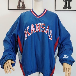 May include: A blue Kansas windbreaker with red and white detailing. The word "KANSAS" is across the chest in white with a red outline. The jacket features a V-neck and elasticated cuffs. The jacket has a drawstring at the hem.