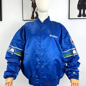 Könnte beinhalten: Blaue Satinjacke mit weißen und grünen Streifen auf den Ärmeln. Auf der Brust steht in weißer Schrift das Wort "Seahawks", und auf dem Ärmel befindet sich ein Teamlogo-Patch. Die Jacke ist zugeknöpft und hat einen gerippten Kragen und Bündchen.