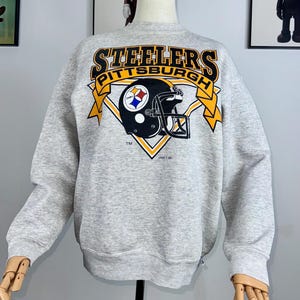 Può includere: Una felpa grigio chiaro con il logo dei Pittsburgh Steelers. Il logo include il nome della squadra in oro e nero, un casco da football nero con un diamante giallo e nero e il nome della città. La felpa ha le maniche lunghe.