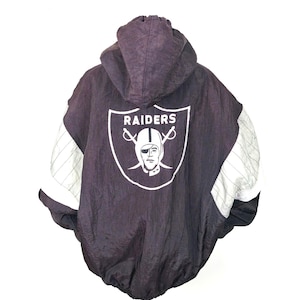 May include: Black and silver Raiders hooded jacket. The jacket features a large Raiders logo on the back, with white lettering and a helmeted face. The sleeves have silver and white striped accents. The jacket is displayed on a metal stand.