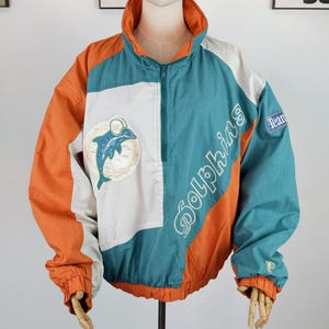 Puede incluir: Chaqueta cortavientos vintage de los Dolphins en color turquesa, naranja y blanco. La chaqueta tiene cierre de media cremallera, cuello alzado y el nombre y el logotipo del equipo. Las mangas y el dobladillo son elásticos.