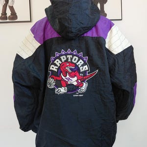 May include: Black, purple, and white Toronto Raptors jacket with a hood. The back features a Raptors logo with a red dinosaur holding a basketball. The jacket has a zip and is made of a shiny material.