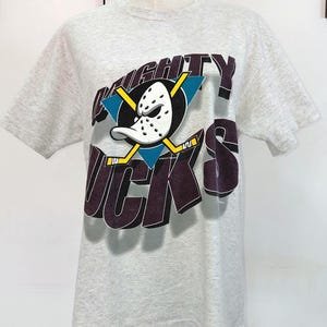 Op de afbeelding: Een lichtgrijs t-shirt met het Mighty Ducks-logo. Het logo bevat de teamnaam, een hockeymasker en gekruiste hockeysticks. Het shirt wordt op een mannequin getoond.