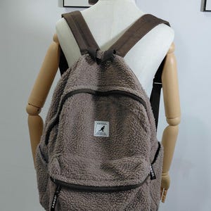 May include: A brown, fluffy backpack with brown straps and a front pocket. The backpack has a zipper closure and a small white tag with the brand name "KANGOL" and a kangaroo logo. The backpack is displayed on a mannequin.