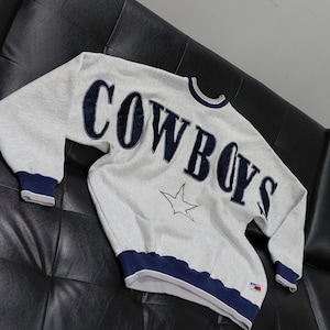 Può includere: Una felpa grigio chiaro con la scritta "COWBOYS" in lettere maiuscole blu navy sul petto. Una grafica a stella bianca è sotto il testo. I polsini, lo scollo e l'orlo sono rifiniti in blu navy.