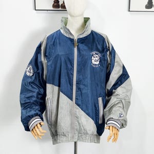 May include: A navy blue and gray zip-up jacket with a geometric design. The jacket features the text "Connecticut Huskies" and a logo on the chest. The sleeves have white and gray striped cuffs.