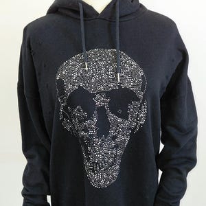 May include: Black hoodie with a large skull design made of small, shiny embellishments. The hoodie has a drawstring hood, long sleeves, and ribbed cuffs and hem. The skull design is centered on the front of the garment.