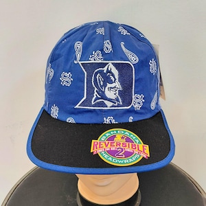 May include: A blue and black reversible baseball cap with the Duke University logo. The cap has a paisley pattern on the blue fabric and a black peak. The tag reads "Reversible 2 times the hat".