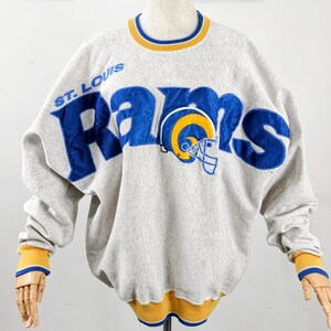 Puede incluir: Sudadera gris con la palabra "RAMS" en azul y un gráfico de casco amarillo y azul. El texto "ST. LOUIS" está encima de la palabra "RAMS". El cuello y los puños son amarillos con ribete azul.