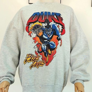 Puede incluir: Sudadera gris claro con un gráfico de un diablo azul encestando un baloncesto. La palabra "DUNKS" está encima de la figura en rojo y azul, y "Blue Devils" debajo en amarillo. La sudadera tiene mangas largas.