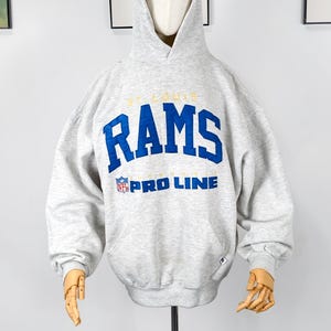 Può includere: Una felpa con cappuccio grigio chiaro con la scritta "ST. LOUIS RAMS" in lettere blu e oro. La felpa con cappuccio presenta anche il logo NFL Pro Line. La felpa con cappuccio è esposta su un manichino.
