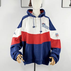Può includere: Una felpa con cappuccio a blocchi di colore bianco, rosso e blu navy, con la scritta "Patriots" in corsivo. La felpa ha un cappuccio con coulisse, una tasca a marsupio e una toppa con il logo NFL sulla manica. L'indumento è esposto su un manichino.