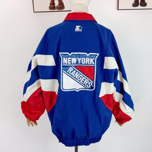 May include: A blue New York Rangers Starter jacket with red and white detailing. The jacket has a large team logo on the back and white stripes on the sleeves. The collar is red, and the Starter logo is visible.