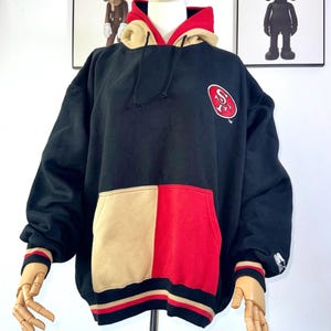 May include: Black hoodie with a red and tan color block pocket and hood lining. The San Francisco 49ers logo is on the chest. The cuffs and hem have red and tan stripes. The hoodie is displayed on a mannequin.
