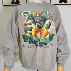 Può includere: Una felpa grigia con una grafica di giocatori di football americano in uniforme verde e gialla. Le parole "PACKERS GREEN BAY" sono stampate in verde. La felpa è esposta su un manichino.
