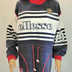 May include: A navy blue, white, and red vintage sweatshirt on a mannequin. The sweatshirt has horizontal stripes, the brand name "ellesse", and the number "19" on the sleeve. It features a drawstring and a hood.