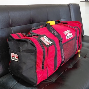 Peut inclure: Sac de sport rouge et noir avec le logo Marlboro. Le sac a une base et des accents noirs, une tirette de fermeture éclair jaune et une bandoulière noire. Le sac est posé sur un canapé en cuir noir.
