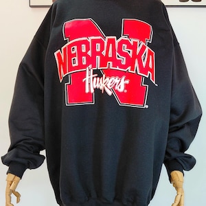 May include: Black sweatshirt with the words "NEBRASKA Huskers" in red with a white outline. The sweatshirt has long sleeves and a crew neck. The design is centered on the chest.