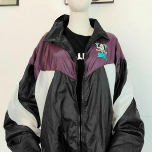 Op de afbeelding: Een vintage windjack in zwart, wit en paars, met een volledige ritssluiting. De jas heeft een turquoise en wit logo op de borst. De jas wordt op een mannequin getoond.