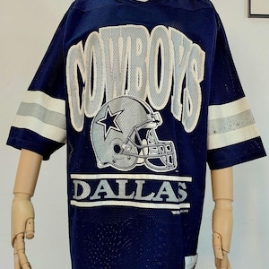 Può includere: Maglia in rete blu navy con la scritta "COWBOYS" in lettere bianche e grigie. Sotto, un casco da football grigio con una stella. La parola "DALLAS" è stampata sotto.