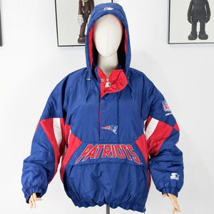 May include: A blue, red, and white Patriots anorak jacket with a hood. The jacket features the Patriots logo and team name across the front. The jacket has a drawstring hood and a front pocket.