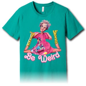 Pode incluir: Camiseta verde-azulado com um gráfico de uma figura semelhante a uma boneca em um vestido rosa e botas amarelas de pele de cobra. A figura está fazendo o espacate. O texto "Be Weird" está em script rosa abaixo.