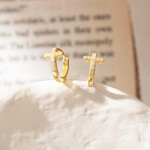 May include: A pair of gold-colored hoop earrings, each featuring a cross design. The crosses are adorned with small, sparkling stones. The earrings are displayed on a white, textured surface, with a blurred book in the background.