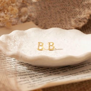 May include: Gold-coloured stud earrings, each in the shape of the letter "B", adorned with small, clear gemstones. The earrings are presented on a white, scalloped dish, placed on a page of printed text.