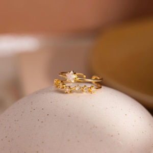 May include: A gold-toned open ring featuring a small star-shaped charm with a pearlescent inlay and a secondary band segment adorned with a row of five tiny, clear, sparkling stones arranged in a delicate, linear pattern.