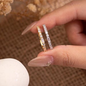 May include: Two delicate rings are held up, one gold and one silver. The gold ring features small, round gemstones and a few rectangular stones. The silver ring has rectangular and round gemstones. The rings are displayed against a neutral background.