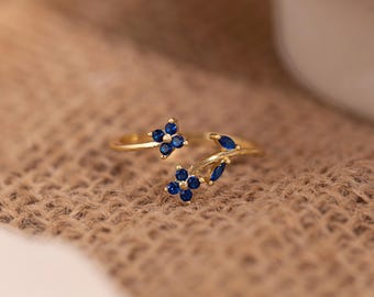 May include: A gold-colored ring with a floral design. The ring features two flower clusters and leaf accents, each adorned with small, round, dark blue gemstones. The ring is set against a textured, tan background.