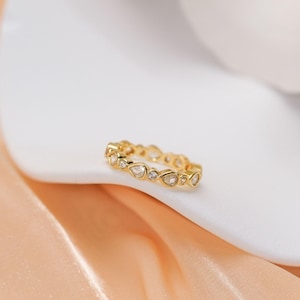May include: A gold band ring featuring a series of alternating pear-shaped and round clear stones set along the entire circumference. The metal has a polished finish, and the stones are arranged in a repeating pattern around the circular band.