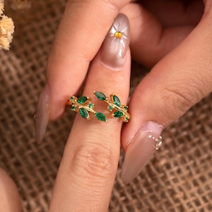 Op de afbeelding: Een goudkleurige ring met een bladvorm, versierd met groene edelstenen. De ring is open, met het bladerenontwerp dat de vinger omsluit. De edelstenen zijn smaragdgroen en ovaal, gezet in een gouden band. De ring is een sieraad.
