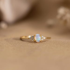 以下が含まれることがあります： 楕円形のオパールを中央に配し、小さなアクセントストーンをあしらった繊細なゴールドリング。 リングは細いバンドと磨かれた仕上げが特徴です。 オパールは乳白色と青色をしています。普段使いに最適です。