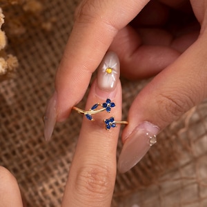 May include: A gold-colored ring featuring a floral design with blue gemstone accents. The ring has a delicate, open design with two flower clusters and leaf details. The ring is shown on a finger against a neutral background.