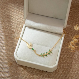 May include: A gold-colored necklace featuring a dove pendant and a branch with green leaf accents. The necklace is displayed inside a white jewelry box, set against a neutral-toned fabric background. The necklace is delicate and elegant.