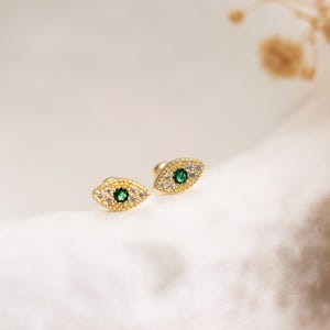 May include: A pair of gold-colored stud earrings, each shaped like an eye. The center of each eye features a green gemstone, surrounded by smaller clear and white gemstones. The earrings are displayed on a white surface.