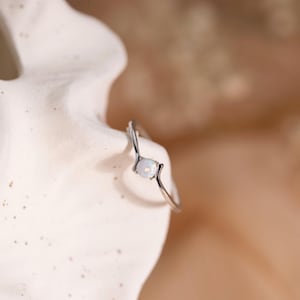Puede incluir: Un anillo de plata con una pequeña gema de ópalo blanco y redondo. El anillo tiene un diseño delicado y curvado, y el ópalo está engastado en un estilo minimalista. El anillo se muestra sobre una superficie blanca texturizada sobre un fondo melocotón suave.