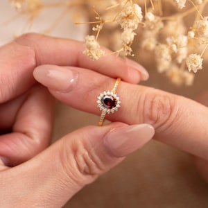 May include: A gold ring featuring a central oval-cut, deep red gemstone surrounded by a halo of smaller, clear gemstones. The ring's band is also adorned with small, sparkling accents. The ring is held up against a soft, neutral background.