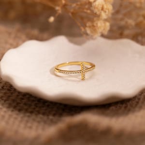 以下が含まれることがあります： 小さな輝く石で飾られた、十字架のデザインのゴールドカラーのリング。リングは、背景にドライフラワーのある、白いスカラップエッジの皿の上に置かれています。リングはジュエリーです。