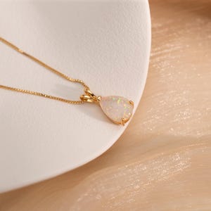 May include: A gold necklace with a pear-shaped opal pendant. The opal has a milky white base with flashes of color. The chain is a delicate gold link. The pendant is set in a gold bezel.